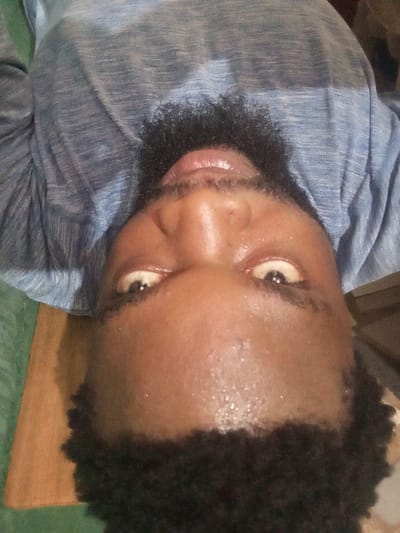 An upside-down, close-up selfie of a dark-skinned man. He has a short, dark beard and curly hair. His eyes are wide and looking towards the camera. His forehead appears sweaty and reflects light. He is wearing a blue and gray mottled shirt. The background includes a wooden surface and a glimpse of green fabric or wall.