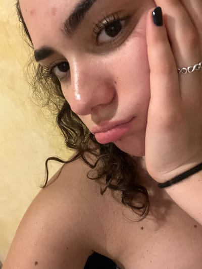 A close-up, tilted image shows a person with dark curly hair and brown eyes. The person's face is partially obscured by their hand, which has dark nail polish and a silver ring. The person's tongue is slightly protruding. The background is a blurry, light beige color. The location, Brisas de Zicatela, Mexico, cannot be determined from this image alone.