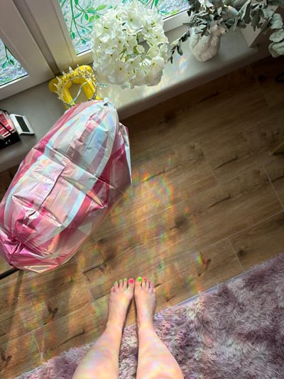 The image depicts a domestic scene, likely indoors, given the presence of a wooden floor and a soft, fluffy rug. Sunlight streams in, creating a rainbow effect on the floor, suggesting a bright, possibly sunny day. The scene is characterized by a collection of objects: a large, foil balloon with pink and white stripes, a bouquet of white flowers, a decorative crown-like object, and a white ceramic sculpture resembling a heart. In the foreground, a person's bare feet are visible, with brightly painted toenails in pink and lime green, resting on the rug. The overall mood is relaxed and casual, with the colorful sunlight adding a playful and cheerful element. The context of Бараўлянскі сельскі Савет, Belarus, places the scene in a specific geographical location.