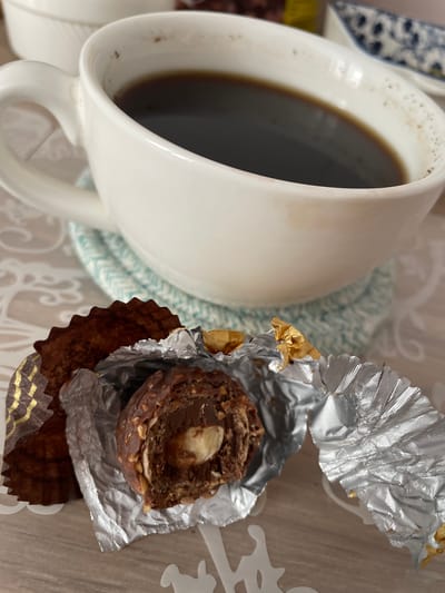 A close-up, overhead shot captures a white ceramic coffee mug filled with dark coffee, placed on a blue and white patterned coaster. To the right of the mug, a partially unwrapped chocolate truffle with a hazelnut center sits in its crinkled silver and gold foil wrapper. The truffle appears to be a Ferrero Rocher. The background is a light-colored, textured surface, possibly a table, with a subtle white lace-like placemat or runner visible. The lighting suggests it is daytime. The scene conveys a sense of a quiet moment, perhaps a break or a treat enjoyed with coffee. No people are visible. The setting appears to be indoors. No visible text is present. The location context is Brovary, Ukraine.