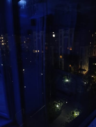 The image depicts a nighttime view from what appears to be a window. Outside, there is a street scene with a multi-story building illuminated by various lights, some from within apartments. Streetlights are also visible in the landscape below, lining a pathway. The interior of the window is dark, with a vertical blue reflection suggesting it may be a curtain. The quality of the picture is dark, and the location (Krykhivtsi, Ukraine) cannot be confirmed from the image alone. The description does not include any subjects or people.