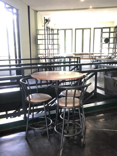 The image shows a small cafe area located on a raised platform, with a wooden table and three black metal bar stools. The table is made of dark wood with a light brown top. The stools are black with brown leather seats. The platform is surrounded by a black metal railing. The floor is made of gray concrete. There is a large window behind the table looking out onto an outdoor area with more tables and chairs. There are also a few shelves behind the table. There is a black ceiling fan on the ceiling. The image was taken in a cafe located at coordinates -7.98 latitude, 112.617 longitude, in the city of Malang, Indonesia.