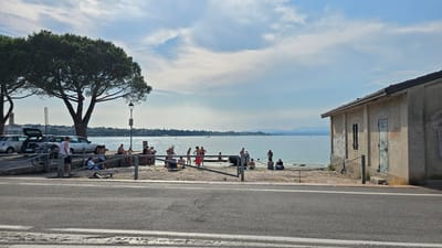 A sunny day at the lakeside in Peschiera del Garda, Italy, as suggested by the distant mountains and the general atmosphere. The scene depicts people enjoying the water and the shore. Several individuals are in the shallow water, some wading and others appearing to swim or cool off. Others are gathered on the stone embankment, either sitting or standing, engaged in conversation or simply relaxing.

In the foreground, a paved road with white markings runs horizontally across the image. Behind the road is a metal railing that separates the walkway from the embankment. To the left, parked cars are visible, including one with its trunk open. A bicycle is also leaning against the railing. A large, mature pine tree with lush green foliage dominates the left side of the image, providing shade. A lamppost stands near the tree.

On the right side of the scene, a small, weathered building with a corrugated roof is situated close to the water. The building's exterior is marked with graffiti, featuring various tags and drawings in different colors. The overall mood is relaxed and leisurely, indicative of a warm summer day at a popular waterfront location. The sky is a mix of blue with scattered white clouds, suggesting fair weather. The time of day appears to be afternoon, given the angle of the sunlight.