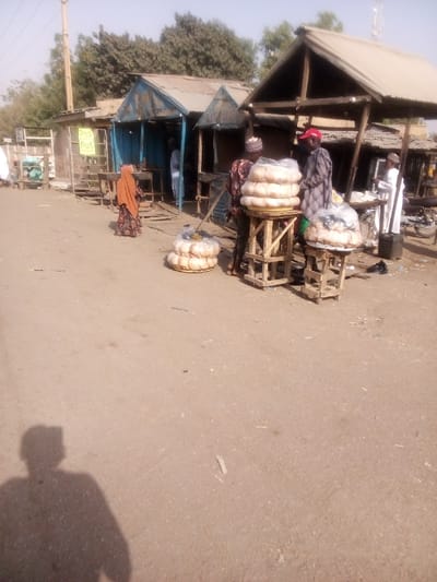 The image depicts an outdoor market or street scene in Dakata, Nigeria, bathed in bright, clear daylight, as evidenced by the distinct shadow of the photographer in the bottom left foreground. The ground is unpaved and appears dusty.
The central focus of the scene is the sale of numerous large, round loaves of bread, likely a local staple. These loaves are neatly stacked on rustic wooden stands of varying heights, and some are also placed on the ground, many covered in clear plastic bags for protection.
Several people are present in the mid-ground. On the left, a person wearing an orange or brown head covering and a patterned dress is seen from behind. Closer to the right, two men in traditional attire, one wearing a red cap, stand near the bread displays, seemingly involved in sales or purchases. Further back on the right, another person in white clothing is visible near a stall that appears to have a motorcycle or bicycle beside it. A person is also partially visible inside one of the background stalls.
In the background, a row of simple market stalls or shops with corrugated metal roofs are visible, some painted blue. A utility pole stands on the far left. A notable detail is a yellow sign hanging from a wooden stand on the left, which clearly displays the text "POS" repeated several times, indicating the availability of Point of Sale services. The scene conveys a typical, active market environment.
