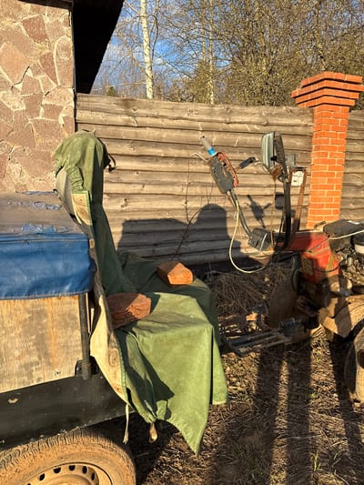 The image shows an outdoor scene in Galevo, Russia, featuring various objects and materials. In the foreground, there's a trailer with a dirty tire visible. A blue and brown object sits on the trailer, covered partially by a green tarp. Several bricks are scattered on top of the tarp. Behind the trailer, there's a motoblock with orange and black elements. The motoblock is parked on dry grass and dirt. A wooden structure, potentially a barn or shed, is visible with a section built from stone. A brick chimney stands atop the wooden structure. A bare tree is visible against a bright sky in the background.