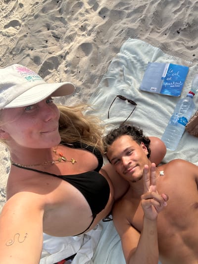 Two people are lying on a white towel on a sandy beach, likely Sainte-Anne, France, as indicated by the water bottle brand visible. A man, with tanned skin and curly dark hair, is lying on his back, displaying a peace sign with his right hand. He wears a silver necklace. A woman, wearing a white cap with a floral design and a black bikini top, is lying next to him, facing towards him. She also wears a necklace and has a tattoo on her left arm. A bottle of water and a book titled "Le ciel est partout" are positioned near the towel. Sunglasses are placed on the towel as well. The scene appears to be a sunny day at the beach.