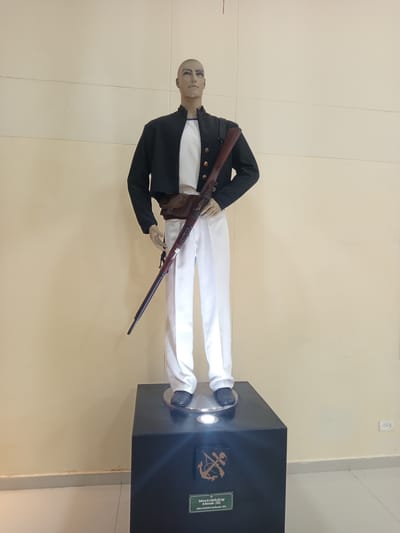 A mannequin dressed in a historical military uniform stands on a dark pedestal in a museum in Coveñas, Colombia. The uniform consists of a black cropped jacket over a white shirt, white trousers, and dark shoes. A brown leather bandolier with a pouch is worn across the chest, and a long rifle with a wooden stock is held diagonally across the body. The background is a plain, light-colored wall. A plaque on the pedestal displays an anchor emblem and text in Spanish and English identifying the uniform as "Uniforme de la Batalla del Lago de Maracaibo - (1823)" or "Uniform of the Battle of Lake Maracaibo - (1823)". The scene is indoors, with soft lighting.