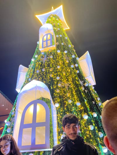 A rotated close-up shot captures a towering Christmas tree adorned with warm yellow lights, white spherical ornaments, and snowflake decorations. Illuminating white house-shaped structures are attached to the tree. Two individuals are partially visible; one at the top corner and another at the bottom. A person is positioned at the right side. The background is a dark night sky.