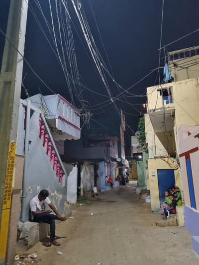 The image shows a nighttime scene in Pedda Kammavaripalli, India. A man sits on a low concrete structure on the right side of the image, looking at a mobile phone. The background is a street lined with multi-story buildings, varying in color. Overhead, a dense network of electrical wires stretches across the sky. A group of children are visible near a blue doorway of a building at the end of the street. There is debris and trash scattered on the street near the man. The overall lighting is dim with light sources from streetlights and buildings.