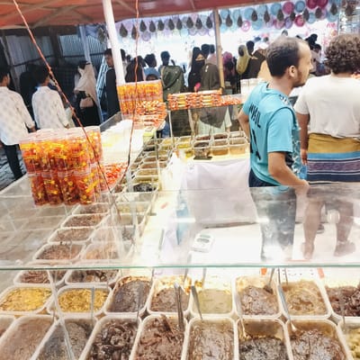 This image captures a vibrant scene at an outdoor market stall, likely in Charfasson, Bangladesh, during the daytime. The foreground is dominated by a long glass display counter showcasing a wide array of food products. Numerous rectangular plastic containers are filled with various types of dark, viscous preserves or pickles, locally known as *achar*, with spoons placed in many of them. Alongside these, stacks of transparent cylindrical containers, some labeled "Natural," hold smaller, brightly colored candies or dried fruit snacks. Additional packaged snacks are also visible on the counter.

In the midground, several people are gathered around the stall. A man in a light blue t-shirt, with the partial word "MRUL" or "MWRUL" visible on his back, stands observing the scene to his right. Next to him, another man with curly hair wears a white t-shirt and a patterned *lungi*, with his back to the camera. Other shoppers, including women wearing *hijabs* and head coverings, are visible in the bustling background, browsing other stalls or moving through the market.

The setting features a red canopy overhead, providing shade, from which decorative items resembling colorful plates or bowls are suspended. Corrugated metal sheets form the walls of nearby temporary structures, characteristic of local markets. The overall atmosphere is lively, with people engaged in selecting and purchasing the various sweet and savory items on display.
