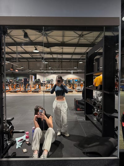The mirror reflects a gymnasium interior with two young women posing. One stands, facing the mirror and holding a phone. The other sits with gym accessories nearby. Behind them are treadmills and various workout machines. The ceiling is exposed, with a metal frame and industrial lighting. Above them in the reflection is a weight rack with dumbbells. The lighting in the gym is bright, highlighting the orange accents of the equipment.