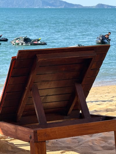 This image captures a scene at Baan Mae Nam, Thailand, likely during the daytime with bright sunlight casting shadows on the sand and wooden structure. The foreground is dominated by a wooden table with a slatted surface, its dark wood showing grain and the construction details like screws. The table is positioned outdoors, with sand dunes and the ocean visible in the background.
In the water, several people are engaged in activities on jet skis. One jet ski is closer to the viewer, adorned with colorful patterns and ridden by at least two individuals. Further out, another jet ski is visible with its rider. The turquoise water appears calm, and the beach is made of light-colored sand. The overall atmosphere suggests a recreational setting, possibly a beach resort or a tourist spot where jet ski rentals are available. The bright sunlight and clear water imply good weather conditions, ideal for water sports. No text is visible in the image.
