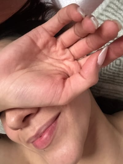 The image presents a close-up, angled view of a person partially covering their face with a hand in what appears to be an indoor setting.
**Subject:** A human individual, gender not definitively discernible but features suggest female.
**People:** One person.
**Actions:** The person's right hand is positioned across their face, obscuring the eyes and forehead. The visible portion of the face, including the nose, mouth, and chin, shows a slight smile.
**Objects:**
* **Hand:** The palm and fingers of a light-skinned right hand are prominent in the foreground. The thumb and index finger display long, pointed, French-tipped artificial nails. The remaining visible fingernails (middle, ring, pinky) appear natural and unpolished. Distinct creases are visible on the palm and knuckles.
* **Face:** The lower portion of the face is visible, including the nose, philtrum, full lips (with a natural pink hue, possibly enhanced with light lip gloss), and chin. Dark hair is visible along the left side of the frame and bordering the face.
* **Environment:** The background consists of a light-colored, textured textile with a subtle geometric or diamond pattern, suggesting a soft surface like a blanket, pillow, or upholstery.
**Interactions:** The hand is directly in contact with the upper part of the face.
**Location:** The specific city of Bogota, Colombia, cannot be determined from the visual information provided in the image. The scene depicts an unidentifiable indoor environment.