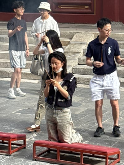 In Dongcheng District, China, on a paved courtyard with stone steps leading up to a red-lacquered building, four individuals are engaged in a ritual involving incense. In the foreground, a woman kneels on a red bench, holding a lit incense stick in each hand and a mask hanging around her neck. To her left, a man wearing a dark blue polo shirt and white shorts holds a single incense stick. Further left and behind him, a man in a white hooded jacket and a bucket hat also holds an incense stick. On the far left, a man in a black t-shirt and grey shorts gestures with his left hand. The scene suggests a religious or ceremonial observance.
