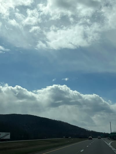 The image shows a view from a vehicle on a highway in the Town of Fishkill, United States of America. The sky takes up the majority of the image, featuring patches of blue sky interspersed with cumulus clouds. A tree-covered mountain is visible along the right side of the image, next to the highway. Cars are traveling on the road. An exit sign is also visible.