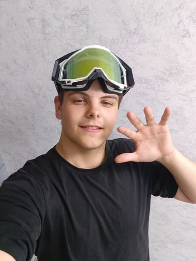 A selfie of a young man wearing black ski goggles on his head. He's wearing a black t-shirt and extending his right hand towards the camera. The background is a mottled gray wall. The picture was taken in the city of Andreyevo, Russia.