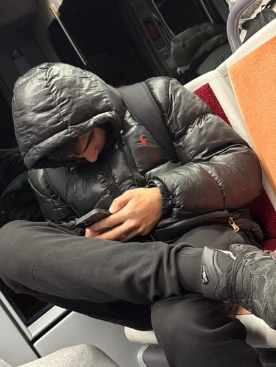 A person sits on a train, wearing a black puffer jacket with the hood up and black tracksuit pants. Their hand holds a smartphone. A pair of grey trainers rests on the jacket. The train's interior features orange seats, black paneling, and white accents. The overall lighting is muted, creating a casual, everyday scene.
