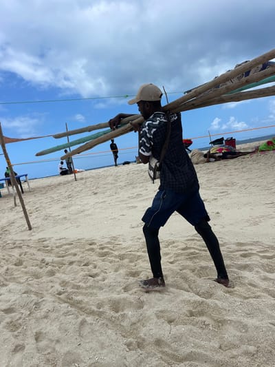 The image displays a sandy beach scene in the city of Zanzibar, Tanzania. A male individual, positioned in the foreground and facing right, is depicted carrying multiple long wooden poles horizontally across his left shoulder and back, stabilizing them with both hands. He wears a light-colored baseball cap, a dark patterned short-sleeved shirt, dark blue shorts over black leggings, and dark footwear. A light-colored shoulder bag with a dark strap hangs from his right side. The ground consists of light-colored sand, marked with footprints. In the middle ground, a dark-clad individual stands upright further left, near the water's edge. To the far left, two other individuals are seated near a light-colored table-like structure. To the right of the primary subject, multiple individuals are resting or lying on the sand, some covered by colorful fabrics. Several vertical wooden poles with horizontal lines (ropes or wires) stretched between them form a visible structure or boundary across the mid-ground. The sky above is partly cloudy with significant blue areas, and the ocean is visible as a distant horizon line.
