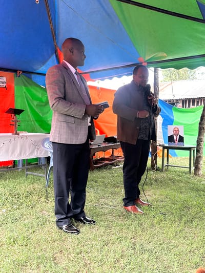 This image, captured in Kiganjo, Kenya, depicts an outdoor public gathering under a large, multi-colored tent. The scene is set on green grass, with natural light suggesting daytime conditions.

Two men are the primary subjects. In the foreground, a man with a bald head stands, dressed in a light grey checkered suit jacket, white shirt, and a striped tie. He holds a smartphone in his right hand and appears to be observing the event. He is wearing dark trousers and black shoes with distinct white soles. Behind him and slightly to the right, another man, wearing a patterned dark shirt, a brown jacket, and dark trousers, holds a microphone, indicating he is either speaking or about to speak. He sports brown, pointed shoes.

Behind the speaker, a banner or poster is displayed on a wooden stand. It features a portrait of an older man with the text "Babu@83," suggesting a celebration, tribute, or political event honoring an individual. Audio equipment, including speakers, is visible near this stand. A partially visible white banner in the lower right corner shows the text "PETER S.C.C." Additionally, a red box in the bottom left displays "L 168/41" and "778971". The overall atmosphere suggests a community event, possibly a political rally or a public meeting, taking place in an open-air setting.