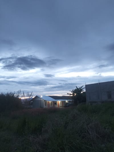 The image depicts an outdoor scene at dusk in Panteraya, Indonesia. The sky is filled with dramatic, dark clouds, suggesting overcast weather or the approach of twilight. In the mid-ground, several structures are visible. To the left, a building with a white roof and orange doors is illuminated by an outdoor light. Adjacent to it, a dark-colored vehicle is parked. To the right of this building, a larger, unfinished concrete structure stands with exposed rebar at the top, indicating ongoing construction. In the foreground, tall, dry grass and vegetation fill the lower portion of the frame, casting the area in shadow and contributing to the somber mood of the scene. A few trees, including what appear to be banana trees, are present between the buildings. The overall impression is one of a quiet, developing rural or semi-rural area during a transitional period of the day. There is no visible text in the image.