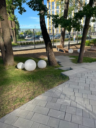 The image shows an outdoor scene with a modern urban aesthetic. In the foreground, a paved walkway made of rectangular grey stones runs diagonally across the frame. To the left of the walkway is a patch of green grass, where three white spherical objects, likely decorative lights or orbs, are placed. Two are larger and one is smaller, arranged in a staggered line.
Several trees with thick, textured bark are visible, with their trunks and branches extending into the frame. Some have lush green foliage. Behind the trees and grass, a black metal fence runs horizontally, separating the immediate area from a more distant scene.
Beyond the fence, a tall, modern building with a yellow and white facade stands prominently, featuring numerous windows. Parked cars and more greenery are visible in the middle ground. The sky is a clear, bright blue, suggesting a sunny day, likely in the late morning or early afternoon given the lighting. The overall impression is a well-maintained urban outdoor space, possibly a courtyard or park area within a residential complex. The location is identified as Kaluga, Russia. No people are explicitly visible, and no text is present in the image.