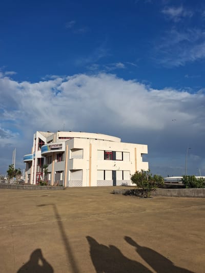 The image depicts a three-story building in Figueira da Foz, Portugal, under a partly cloudy sky. The building is white and gray, with red trim around some windows. Blue and white signage is visible on the balconies. A concrete sidewalk runs along the side of the building, casting shadows from a person or object. A road runs alongside the sidewalk. The sky transitions from a deep blue on the left to clouds and a lighter blue on the right.