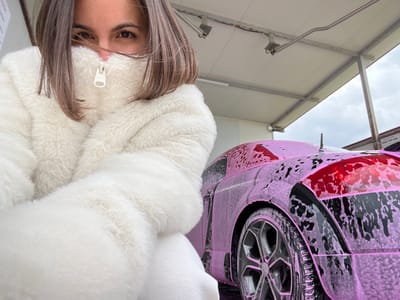 In the city of Bryastovo, Bulgaria, an outdoor car wash bay is depicted under an overcast sky. A female subject, with shoulder-length brown hair and brown eyes, occupies the left half of the frame, looking directly at the camera. She wears a cream-colored, high-collared faux fur jacket, zipped to conceal her mouth and nose, revealing only her eyes and forehead. Her arms are bent at the elbows, holding the jacket around her. To the right, a dark-colored sports car is visible, covered entirely in vibrant pink foam, indicating an active washing process. The car's rear quarter panel, a multi-spoke wheel, and a portion of its roof are discernible through the foam. The car wash structure features a white panel ceiling supported by grey metal beams. In the distant background, to the far right, a building wall with a partially visible sign displaying the phone number "0882 750 436" can be observed.