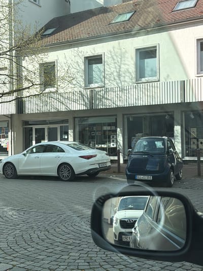 The image shows a street scene in Ulm, Germany. A white Mercedes-Benz CLA coupe is parked on the cobblestone street, parallel to a building with storefronts and apartments above. A blue electric car, a Microlino, is parked behind it. The reflection in the side mirror of the car taking the picture shows a white Hyundai SUV. The buildings have windows and awnings. A tree with budding leaves is visible on the left side of the frame.