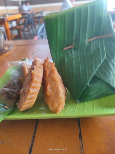 In Geulumpangpayong, Indonesia, a close-up shot captures a bright green rectangular plate on a wooden table. The plate holds two golden-brown fried snacks, possibly pisang goreng (fried bananas), served alongside a package wrapped in a vibrant green banana leaf, secured with small wooden skewers. In the background, the scene is blurred, revealing the interior of a restaurant with wooden furniture and indistinct figures of people seated at tables. The overall lighting suggests a daytime setting.