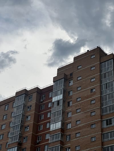 This media file shows a close-up, tilted view of a modern, multi-story brick building with a large portion of cloudy sky. The building's facade is characterized by rows of small, rectangular windows and some larger, multi-pane windows that appear to be balconies or sunrooms. The brickwork is a reddish-brown color, and vertical concrete or stone strips divide sections of the brick. There are also visible air conditioning units attached to some parts of the building. The sky is filled with heavy, gray clouds, suggesting overcast or stormy weather. The lighting indicates it is daytime, but the cloudy conditions create a diffused, somewhat somber atmosphere. The image is oriented at an angle, making it difficult to discern a specific location within Irkutsk, Russia, beyond it being an urban residential or commercial building. No people are visible, and the focus is entirely on the architecture and the sky. The jagged line of the building's edge against the sky is a notable visual element.