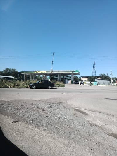 A black sedan is driving past a gas station with a green canopy and the logo "O KARAMAI". The gas station appears to be located in Talgar, Kazakhstan. The sky is clear and blue. The road is paved, and there are some bushes and trees in the background. There are also some other vehicles parked near the gas station and a tall transmission tower in the distance. The overall scene depicts a typical day at a gas station in a suburban or rural area.