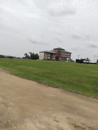 The image shows a large, multi-story building complex located in Uyo, Nigeria, viewed from a ground-level perspective on a dirt road. The building has a distinct architectural style with light-colored walls, brown brick accents, and a green multi-tiered roof. A grassy lawn spans the area between the road and the building, with several cars parked in front. To the right of the building is a large, dark-colored dump truck with an attached flatbed trailer parked on the grass. Utility poles and streetlights are visible near the truck. In the background, some trees and another building are visible in the distance. The sky is overcast with gray clouds.