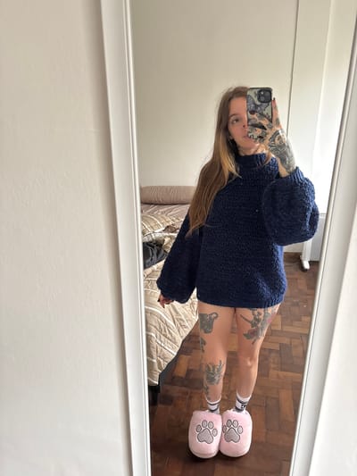 A mirror selfie captures a woman standing in an indoor setting. The woman has long, straight brown hair and extensive tattoos covering her arms and legs, featuring intricate designs. She is wearing a dark blue, oversized chunky knit sweater that appears to be quite plush. On her feet, she sports white ankle socks with black stripes, and soft, pink, paw-print shaped slippers. She holds a smartphone in her right hand, its screen facing away, revealing a phone case adorned with a butterfly pattern. Her gaze is directed towards the camera's reflection in the mirror.
The background reflected in the mirror shows a room with light-colored walls or baseboards in the upper and lower sections. The floor is made of wood, arranged in a distinctive herringbone or parquet pattern. A portion of a bed, covered with a light-colored, quilted blanket featuring parallel stripes, is visible behind her legs in the reflection. The mirror itself has a simple white frame, framing the scene.
The image suggests a casual and comfortable atmosphere, typical of a home environment in Cordoba, Argentina. No specific time of day or weather conditions can be determined from the indoor setting. There is no visible text present in the image.