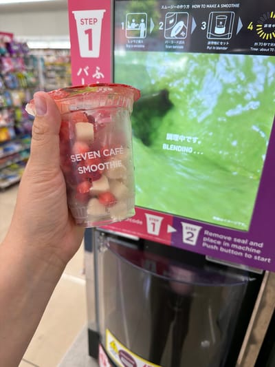 A person's hand holds a clear plastic cup filled with cut strawberries and what appear to be cubes of banana or taro, a popular ingredient in Japanese desserts. The cup is branded "SEVEN CAFE SMOOTHIE." The background features a digital screen displaying instructions for making a smoothie in Japanese and English, with steps labeled "STEP 1," "STEP 2," "STEP 3," and "STEP 4." The screen shows graphics of a person, a barcode, a blender, and a timer. The text on the screen indicates the process involves scanning a barcode, placing the cup in a blender, and the smoothie will be ready in approximately 70 seconds. The environment appears to be a convenience store, likely a Seven-Eleven, given the branding, with shelves of products visible in the blurred background. The purple signage and the smoothie machine suggest a location in Chuo, Japan, a major commercial and business district within Tokyo. The image captures a moment of preparing a drink from a self-serve smoothie machine.
