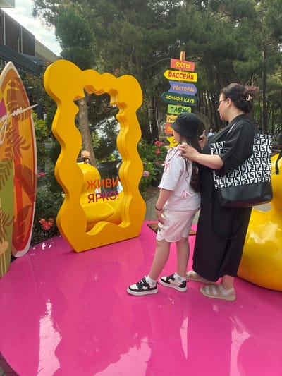 The image depicts a vibrant outdoor scene in Gelendzhik, Russia. In the foreground, a bright pink, glossy circular platform serves as the base for several elements. To the left, a portion of a surfboard with a tropical design is visible. Center stage, a large, bright yellow, abstract, wavy frame stands prominently. Inside this frame, a mirror reflects a yellow, duck-shaped object and text that reads "ЖИВИ ЯРКО!" (Live Brightly!).

Two individuals are interacting in front of the frame. A woman, wearing a black dress and carrying a black and white patterned tote bag, stands on the right. Her right hand rests on the shoulder of a young person in front of her, who is wearing a white t-shirt with pink ribbons attached, white shorts, and a black bucket hat. They are both facing the yellow frame.

In the background, a directional signpost with multiple colorful arrows points to various locations, including "ЯХТЫ" (Yachts), "БАССЕЙН" (Pool), "РЕСТОРАН" (Restaurant), "НОЧНОЙ КЛУБ" (Nightclub), and "ПЛЯЖ" (Beach). Beyond the signpost, tall evergreen trees form a dense forest, suggesting a park-like or recreational area. The sky above is partially cloudy, with patches of blue visible. The overall impression is a lively and colorful attraction, likely a photo opportunity, within a scenic outdoor setting.