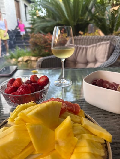 The scene depicts a table laden with various refreshments, likely situated in Salou, Spain. On the table, there are dishes containing pineapple slices, strawberries, grapefruit slices, and salami. A glass of white wine is also present. In the background, there is a view of people near a pool. There is wicker furniture.