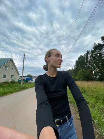 A first-person perspective shot shows a person with blonde hair pulled back in a bun, wearing a black long-sleeved shirt and blue jeans with a gold belt buckle, standing on a dirt road in a rural area. The person is looking to the right, with their left arm and hand visible in the foreground. To the left of the road is a light-colored house with a green roof, surrounded by greenery. A utility pole with power lines is visible between the house and the road. In the background, there is a dense forest of tall pine trees and a field of tall grass. The sky is overcast with grey clouds. The location is identified as Podshivalovo, Russia.