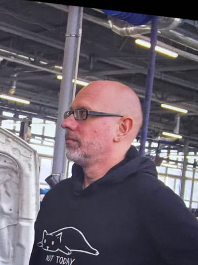 A medium shot captures a bald man with glasses and a goatee, seen in profile from the chest up, standing in an indoor industrial setting. He is wearing a black hooded sweatshirt with a white graphic of a cat and the text "NOT TODAY". The man's left side is prominent, and he is looking to his right. Behind him, the background consists of structural beams, pipes, and fluorescent lighting fixtures, suggesting a workshop or factory environment. Part of a white vehicle or piece of equipment is visible on the left side of the frame.