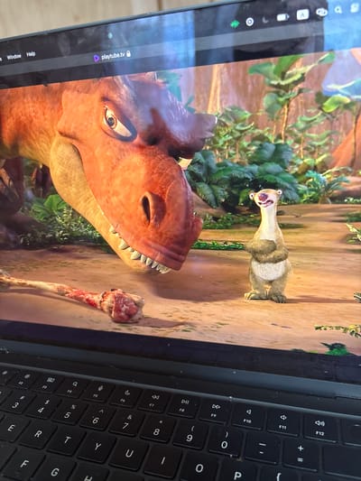 This is a close-up, eye-level, indoor shot of a laptop screen displaying a scene from the animated movie "Ice Age: Dawn of the Dinosaurs." The main subjects are a large, reddish-brown Tyrannosaurus Rex and a small, light-brown sloth named Sid. The Tyrannosaurus Rex is on the left, with its head lowered towards a bloody bone on the ground. Sid is on the right, standing upright with his arms crossed and a wide smile. The background is a lush, green jungle environment. The laptop keyboard is visible at the bottom of the frame, suggesting the image was taken by someone using the laptop. The top of the screen shows a browser tab with the URL "playtube.tv". The location of Anton, Bulgaria is not discernible from the image.