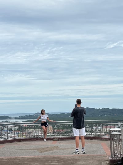 In the city of Ratsada, Thailand, two individuals are positioned on a viewing platform overlooking a sprawling urban landscape and a distant body of water. The platform is bordered by a metal railing and a patterned concrete floor. In the foreground, a woman with long, light brown hair poses with her left hand on a railing and her right arm extended slightly backward. She is wearing a white crop top with black trim and black shorts with white accents. In the mid-ground, a man stands with his back to the camera, holding a smartphone in both hands, seemingly taking a photograph. He is dressed in a dark t-shirt and white shorts, with athletic shoes and socks. The background reveals a densely populated city with buildings of varying heights, interspersed with green foliage. Beyond the city, a calm sea stretches to the horizon, where rolling, tree-covered hills create a scenic backdrop under a cloudy sky.