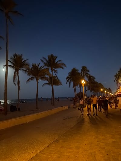 This is a nighttime photograph of the Hollywood Beach Boardwalk in Hollywood, Florida, USA. The scene captures a lively atmosphere with a clear, dark blue sky overhead. On the left, a sandy beach stretches towards the ocean, where gentle waves are visible. Several palm trees are scattered across the beach and along the boardwalk, with their fronds silhouetted against the night sky. Some people are scattered on the beach, seemingly relaxing or walking.

The boardwalk itself, paved with brick-like stones, is illuminated by warm, yellow streetlights mounted on poles. A crowd of people is walking along the boardwalk, appearing to be socializing and enjoying the evening. The individuals are dressed in casual summer attire, suggesting warm weather. Some are walking in groups, while others are walking individually. The overall impression is one of a popular tourist destination at night.