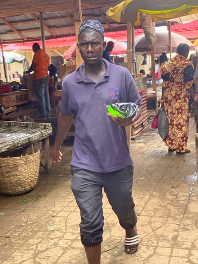 The image depicts a man standing in what appears to be an outdoor market in Kafanchan B, Nigeria. He is wearing a purple t-shirt with an emblem on the left side, dark pants, and striped sandals. He is holding a plastic bag containing a green substance.
The market setting includes stalls with corrugated metal roofs, various items for sale, and other people in the background. The ground appears to be a paved surface. The man is looking at the camera. There are people walking and selling items.