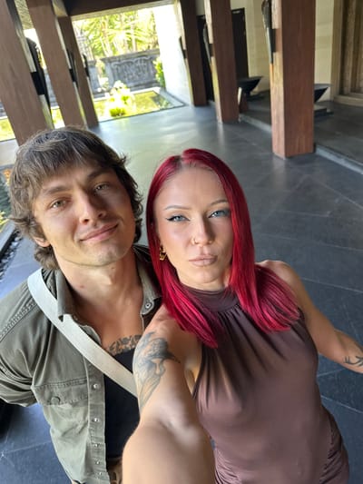 A selfie captures a man and a woman in Nusa Dua, Indonesia. The man, positioned at the bottom of the frame, has light brown hair and green eyes. He is wearing a green shirt with a white strap. The woman, situated above him, has red hair and blue eyes. She is wearing a brown dress. A tattoo is visible on her arm. The background features a blurred view of a building with wooden beams and greenery outside the window. The floor is tiled in dark grey.