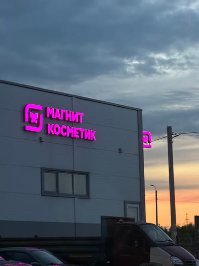 This media file captures an exterior view of a building with prominent signage. The main subject is a building with a gray, corrugated metal facade. A large, illuminated pink sign displaying "МАГНИТ КОСМЕТИК" is mounted on the building. Below the text is the Magnit Kosmetik logo, also in pink, which features a stylized flower or butterfly within a square.
Above the building, a sky is visible, characterized by dramatic clouds and a warm, orange glow from what appears to be a sunset or sunrise. A power line and pole are also visible against the sky. To the right of the building, part of a dark-colored vehicle is visible, with a sliver of another vehicle behind it.
The scene suggests an outdoor setting, possibly in an urban or suburban area, given the presence of the building, signage, and vehicles. The time of day is clearly indicated by the atmospheric lighting in the sky, suggesting either early morning or late evening. The weather appears to be cloudy, contributing to the dramatic sky colors. No people are visible in the media file.