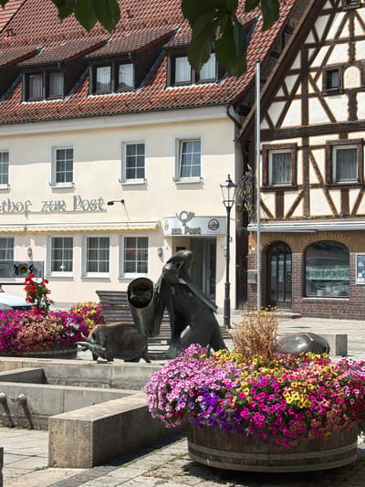 The image depicts a vibrant scene in the Bavarian town of Ebermannstadt, Germany, focused on a public fountain area decorated with abundant, colorful flowers. In the foreground, a large wooden barrel overflows with a dense arrangement of pink, purple, and yellow petunias. To the left of the barrel, a stone basin of the fountain is visible, with two cylindrical spouts protruding from a concrete wall.
Behind the fountain, a bronze sculpture features a bull-like figure carrying a large, cylindrical object on its back, with another smaller bull figure in front of it. A wooden bench is situated behind the sculpture.
The background reveals the architecture of Ebermannstadt. To the left, a light-colored building has a sign that reads "ethor zur Post" and "zur Post," indicating a post office or hotel. This building has multiple windows with white frames and visible curtains. Adjacent to it, a traditional German half-timbered building stands out with its dark brown wooden beams and white plaster infill. This building has arched windows and brickwork at its base, with a sign above the windows that reads "Delzbech Lebensmittel" (groceries). A traditional black lamppost stands between the two buildings.
The overall scene is bright and sunny, suggesting a pleasant day, with green leaves from an overhanging tree visible in the upper left corner of the image. The ground is paved with cobblestones and flagstones. No people are present in the image.