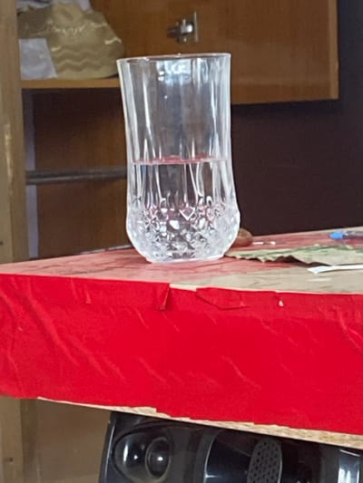 The scene, located in Odo Ona, Nigeria, presents a clear, cut-glass tumbler approximately half-filled with a clear liquid, likely water. The tumbler, featuring a faceted base and smooth upper walls, rests on a light-colored, patterned surface. This surface is partially draped with a vibrant red fabric exhibiting folds and creases. In the background, a blurred wooden cabinet or shelving unit is visible. The cabinet consists of a lighter brown section to the left, displaying a horizontal bar and a shelf holding a light-colored object with a wavy gold pattern. To the right, a darker brown panel with a metallic fixture is visible. Below the table, a dark electronic device with circular elements and a grille is partially visible beneath the red fabric. To the right of the tumbler on the table surface, a small dark object, several white specks of debris, a piece of green-patterned material, and the blue cap of an unknown object are present.