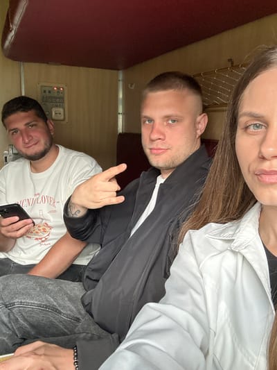 Three individuals are visible in the image, taken at an upward angle, presumably in the interior of a train car, based on the presence of a red bench seat and control panel. The individual at the top, partially visible, is wearing a white shirt. Below and to the left, a second individual is wearing a black jacket over a white shirt and is making a "rock on" hand gesture. The third individual, positioned below and to the left of the second, is wearing a white t-shirt with a graphic design and holding a black smartphone. The image lacks location-specific details needed to confirm the city of Красненскі сельскі Савет, Belarus.