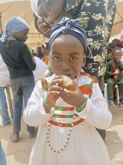 The image depicts an outdoor gathering in Masvingo, Zimbabwe, under a beige canopy, on unpaved, sandy ground. The central subject is a young, dark-skinned child wearing a dark blue knotted headwrap, a long-sleeved white dress adorned with horizontal bands of colorful geometric and striped fabric (orange, green, red patterns) across the chest and waist, multiple green beaded bracelets on each wrist, and a white and red beaded necklace. The child holds a light-brown, textured food item to its mouth with both hands, mid-consumption, and looks directly forward with a neutral expression and prominent lighter-colored eyes. In the mid-ground to the left, another young individual in a light blue headscarf, dark top, and blue jeans stands facing away. To the right, partially obscured, a person in a dark garment with a light floral pattern has hands visible above the central child's head, potentially holding an obscured infant. Further right, a small child in a green outfit and red beaded necklace is seated, also eating.