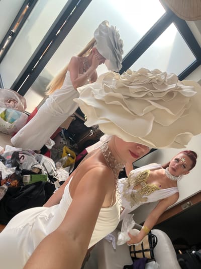The image captures a backstage scene, possibly before or after a performance, with three individuals. Two are prominently featured in the foreground and mid-ground, wearing elaborate white gowns and large floral headpieces. The person in the immediate foreground is partially obscured by a large, creamy white, ruffled headpiece, and is wearing a sparkling necklace and a white dress. Another person, in the mid-ground, is also adorned in a white gown and a sparkling necklace, with intricate gold detailing on the bodice. This individual has striking makeup with facial gems and their hair is pulled back in a red ponytail. They are looking towards the camera.
In the background, another person is visible, also in a white dress, facing away from the camera and appearing to adjust their headpiece. The setting appears to be indoors, with large windows allowing natural light to flood the space, suggesting it could be daytime. The floor is cluttered with various objects, including bags, clothing, and what appear to be props or costumes, indicating a busy, behind-the-scenes environment. The overall mood is one of preparation or winding down from an event, with a focus on the elaborate costumes and makeup. No visible text is present in the image.