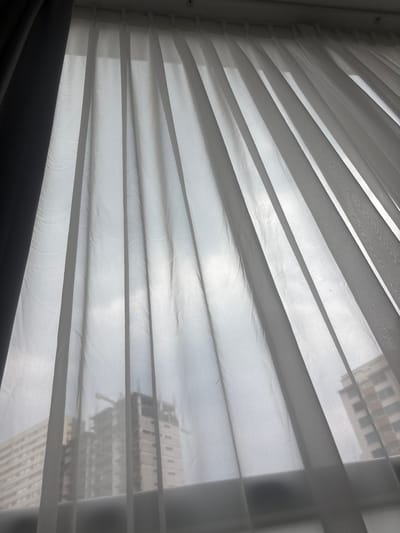 The image depicts a view through a window with partially closed white blinds. Through the blinds, a building can be faintly seen. The blinds are angled, creating a series of horizontal lines and shadows. The overall light level appears bright, possibly due to daylight. The location, Jesús del Monte, Mexico, is not discernible from the image alone.